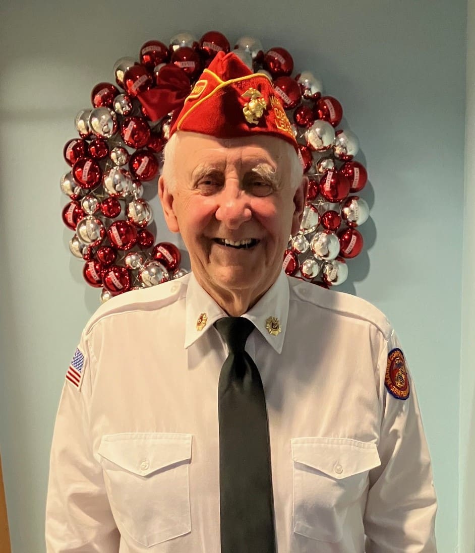 Officer of Marine Corps League with white button up shirt, black tie, and red hat with a holiday wreath behind him.