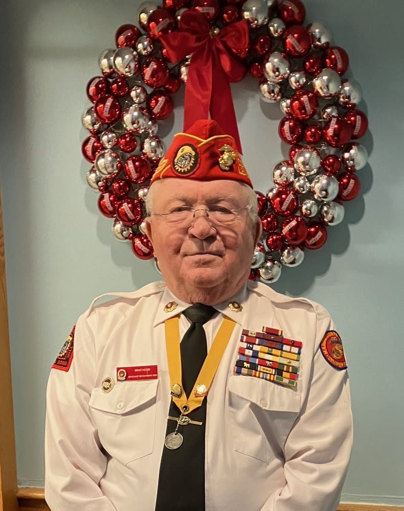 Officer of Marine Corps League with white button up shirt, black tie, and red hat with a holiday wreath behind him.