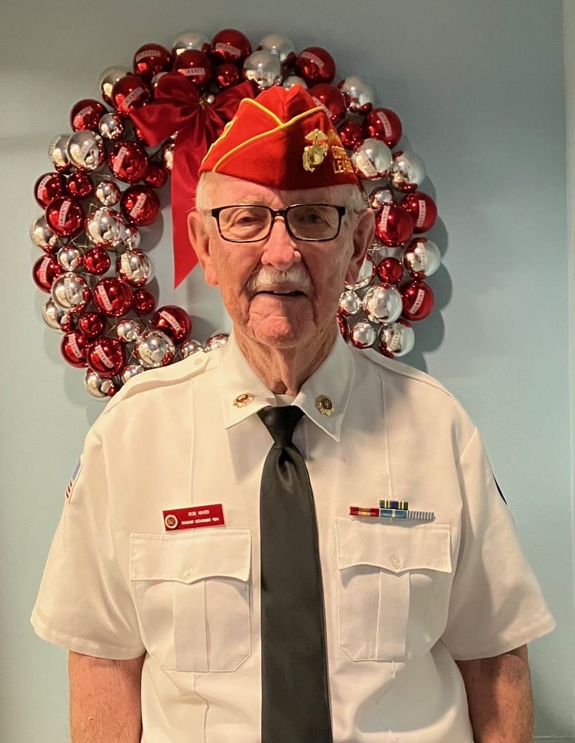 Officer of Marine Corps League with white button up shirt, black tie, and red hat with a holiday wreath behind him.