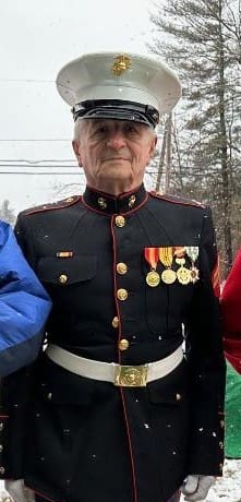 Officer of Marine Corps League with white button up shirt, black tie, and red hat with a holiday wreath behind him.