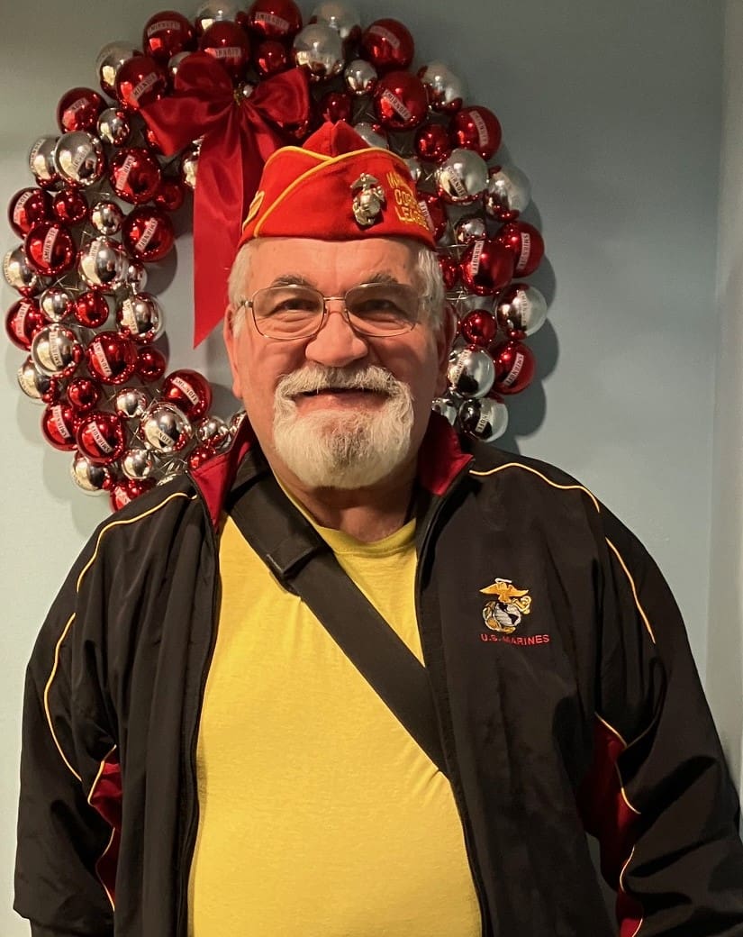 Officer of Marine Corps League with white button up shirt, black tie, and red hat with a holiday wreath behind him.
