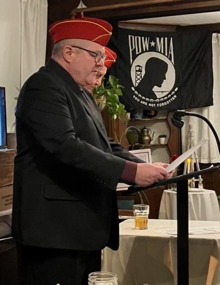 Officer of Marine Corps League with white button up shirt, black tie, and red hat with a holiday wreath behind him.