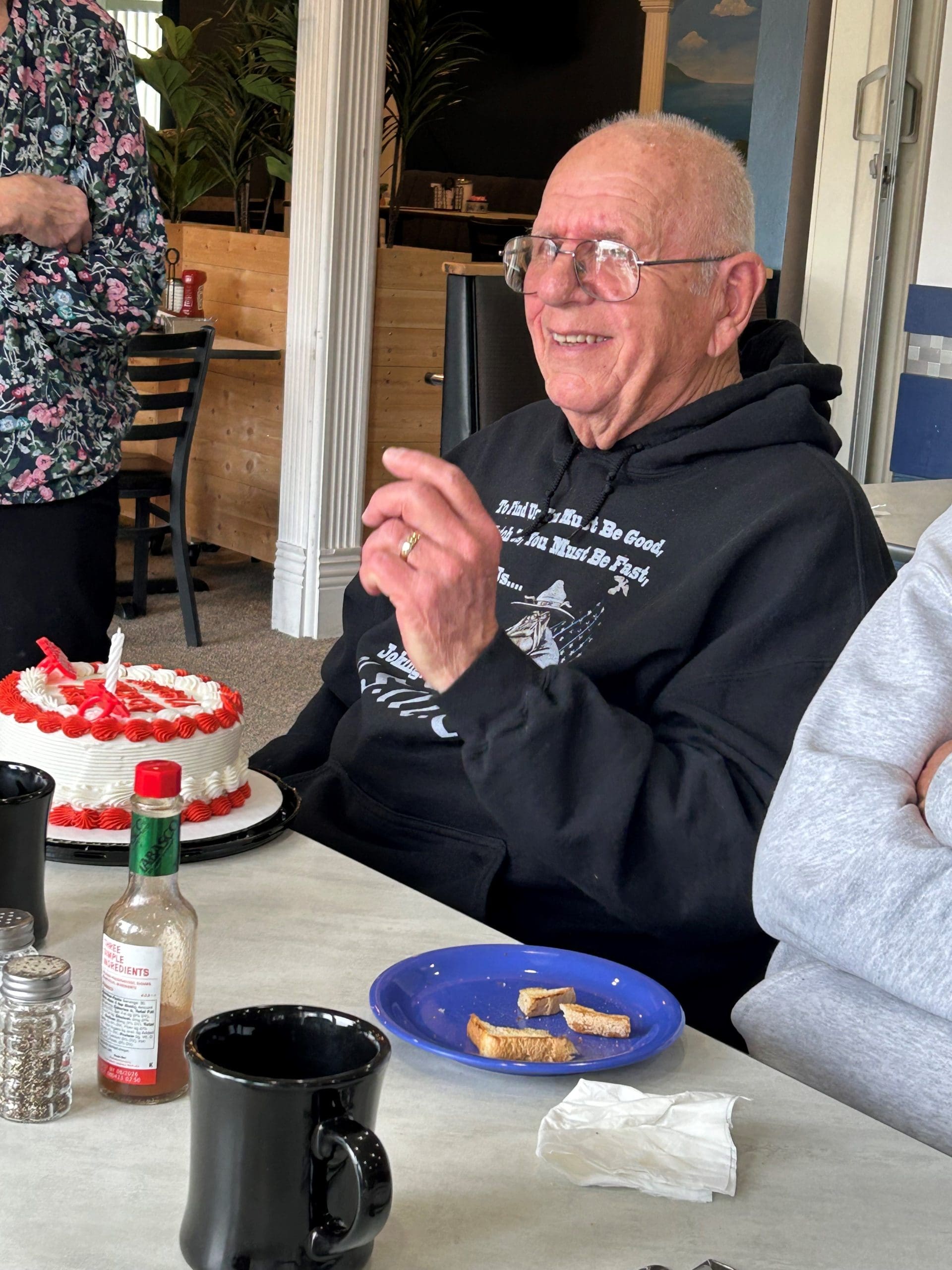 86 year old man with glasses and dark blue sweatshirt celebrates his birthday with a white and red cake.